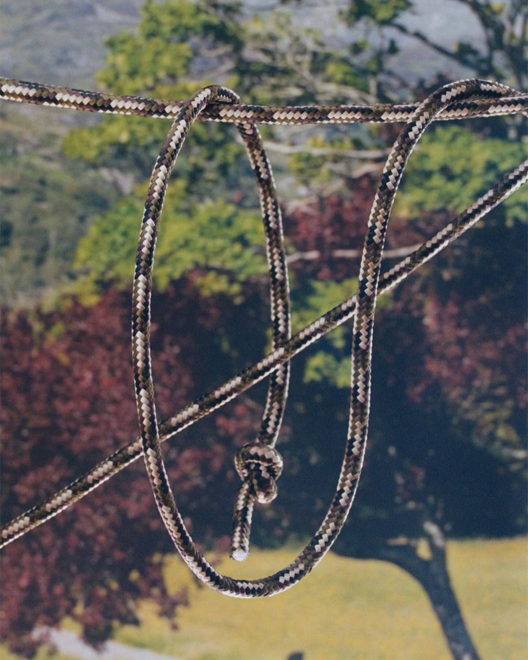 Camouflage rope with a knot is suspended in front of an outdoor background with trees and foliage in a natural light setting.