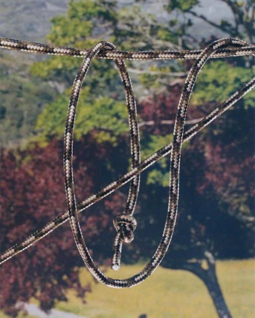Camouflage rope with a knot is suspended in front of an outdoor background with trees and foliage in a natural light setting.