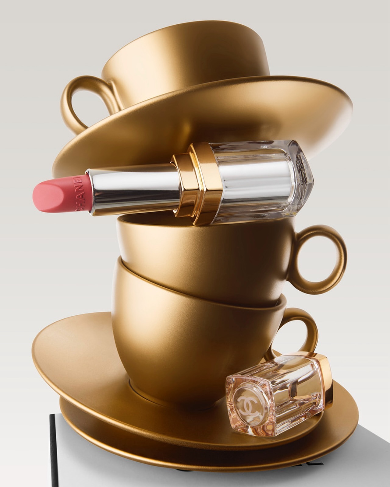 Beauty still life featuring a tower of gold teacups with lipstick and perfume bottle, capturing luxury aesthetic on a white backdrop.