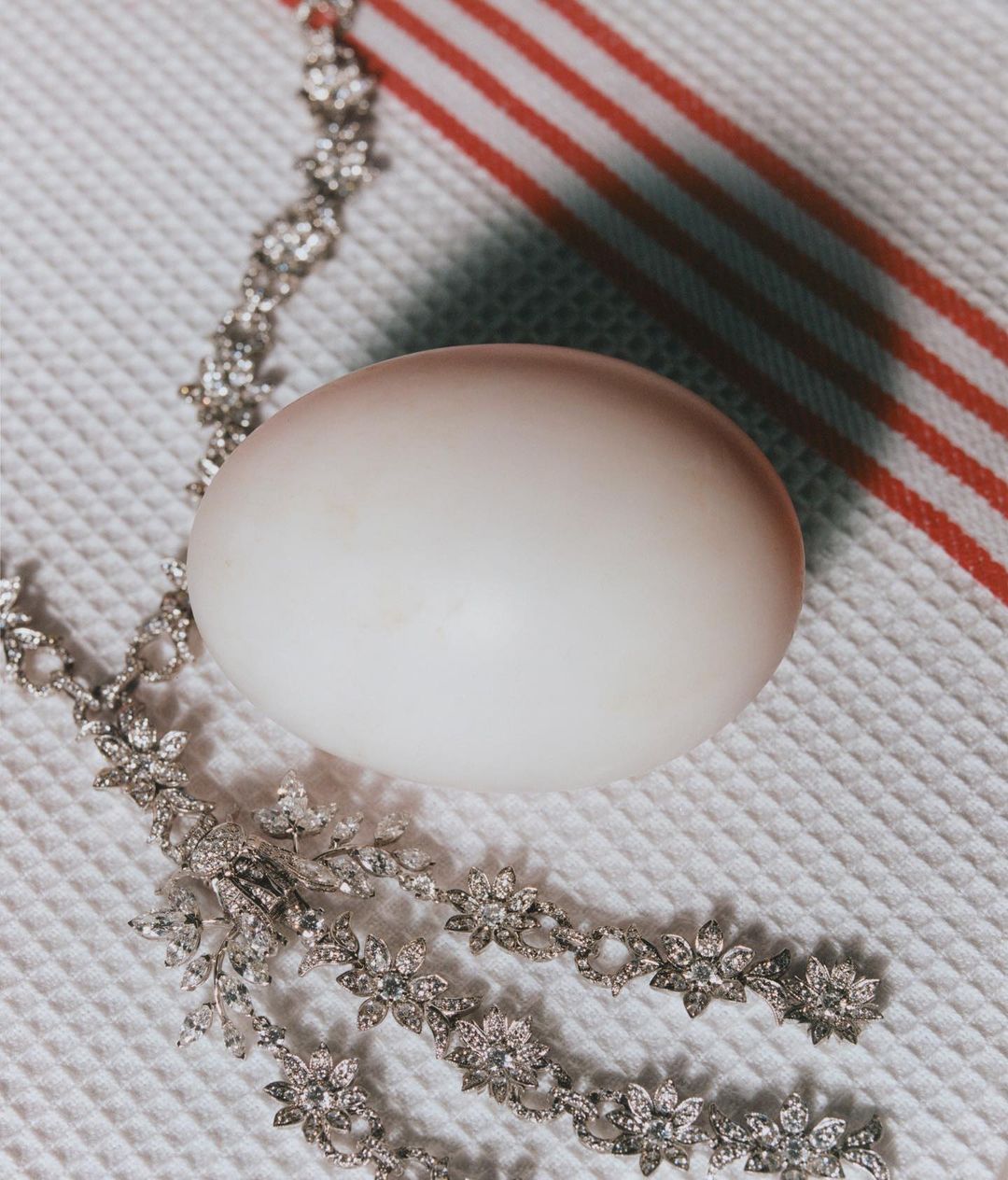Still life of a whole egg with a diamond-encrusted floral necklace on a textured white surface with red stripes in the background.