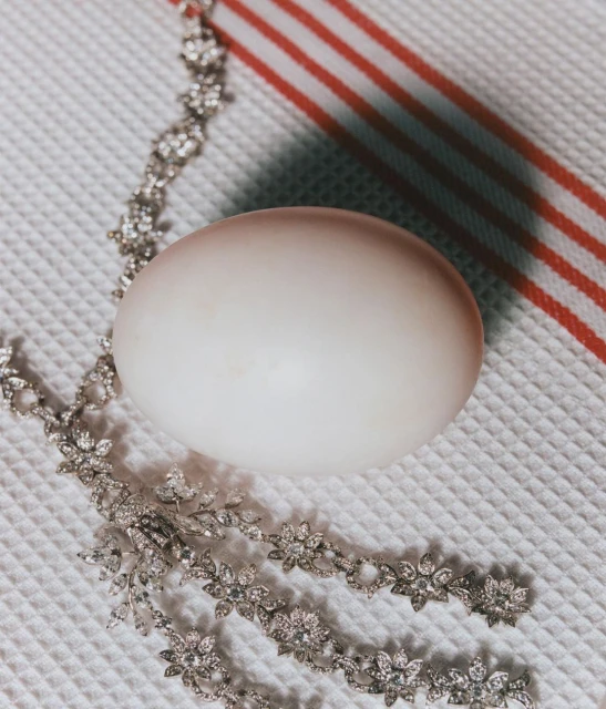 Still life of a whole egg with a diamond-encrusted floral necklace on a textured white surface with red stripes in the background.