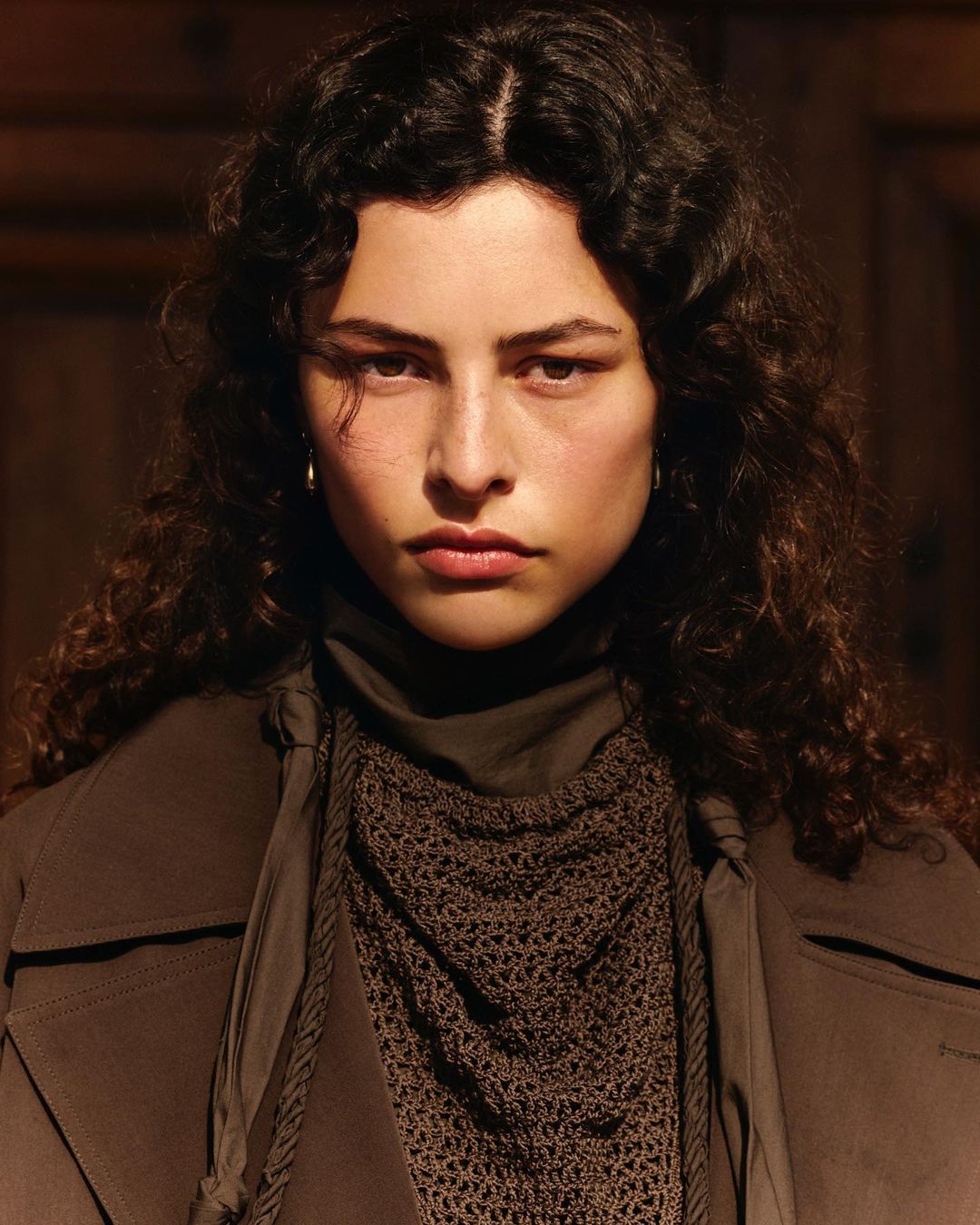 Close-up shot of a woman with curly hair wearing a brown coat and brown scarf, gazing directly at the camera in a dimly lit indoor setting.