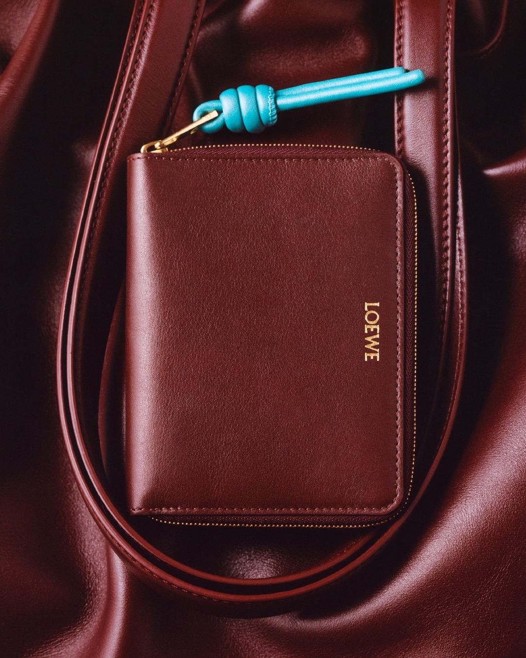 Burgundy leather wallet with gold Loewe logo and blue accent on a burgundy draped leather handbag strap; luxury fashion accessories.