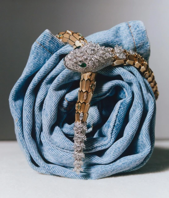 Close-up of a snake-shaped bracelet with gold and diamond detailing coiled around a blue denim cloth in a studio setting.