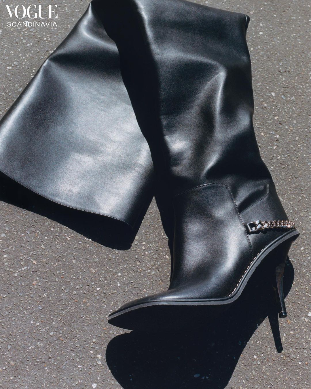 Fashion shot of a single, high-heeled black leather thigh boot with chain detail, displayed on a neutral gray textured surface.