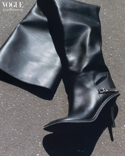 Fashion shot of a single, high-heeled black leather thigh boot with chain detail, displayed on a neutral gray textured surface.