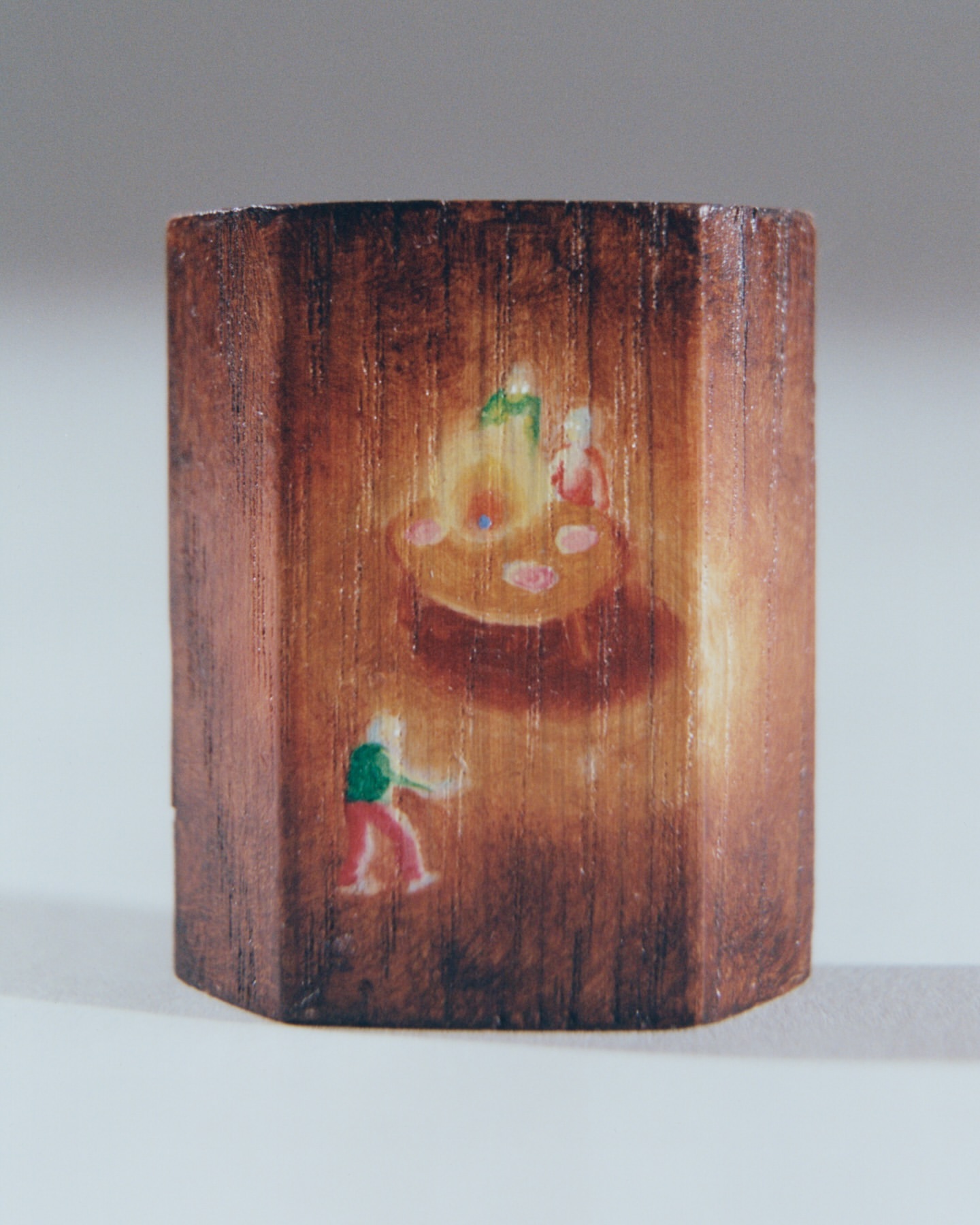 Miniature painted wood block featuring figures around a fire with a standing figure to the side on a light background.