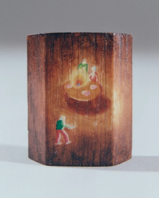 Miniature painted wood block featuring figures around a fire with a standing figure to the side on a light background.