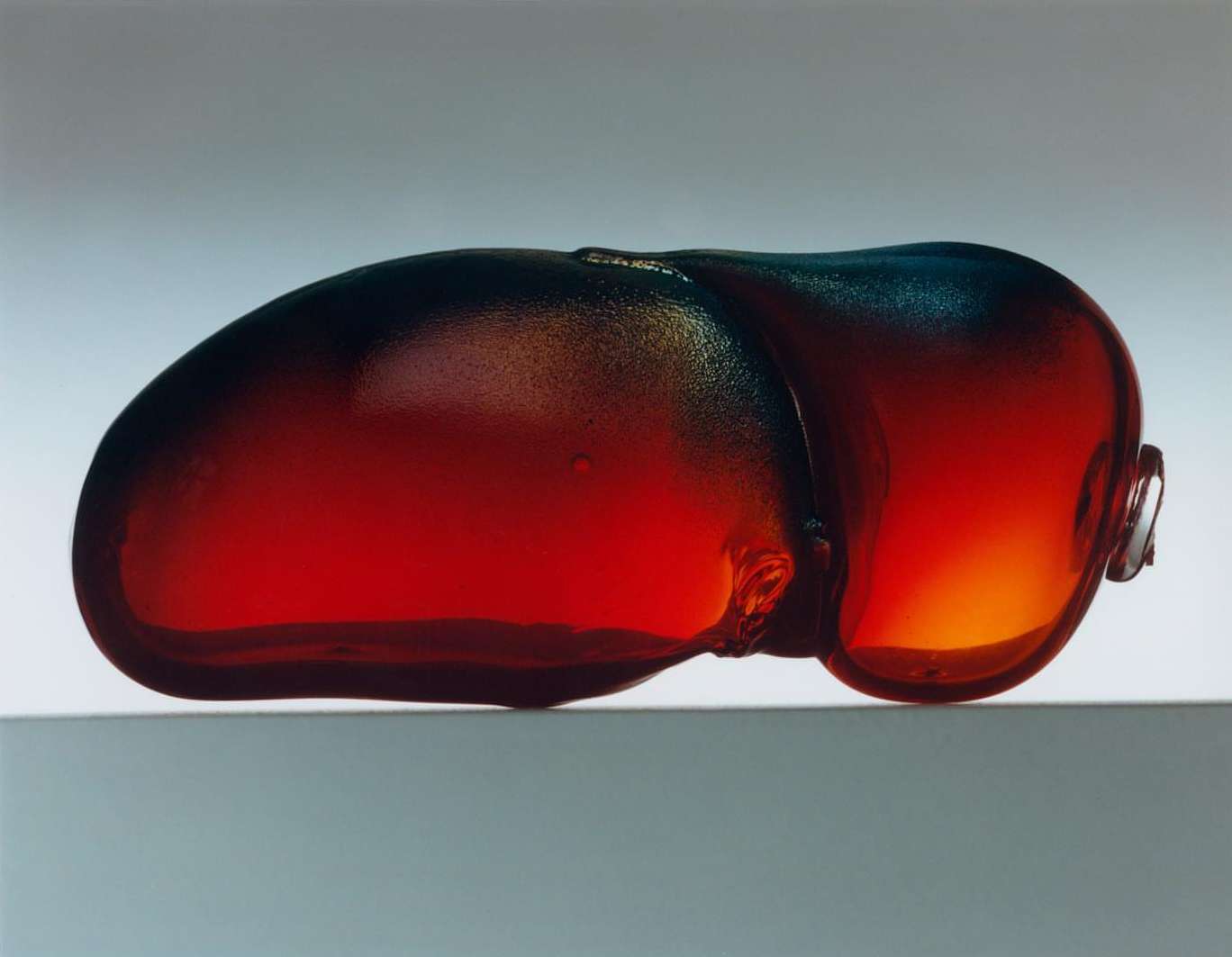Amber-colored glass art piece resting on a surface, showcasing unique form and gradient hues from deep red to translucent amber tones.