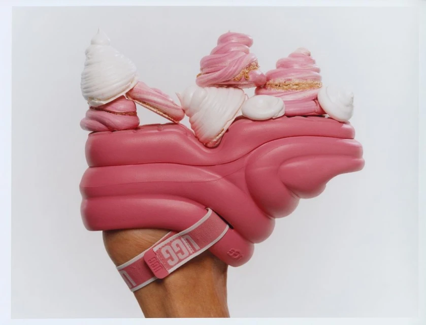 An arm holds a pink platform shoe topped with white and pink imitation ice cream and gold glitter against a white background.