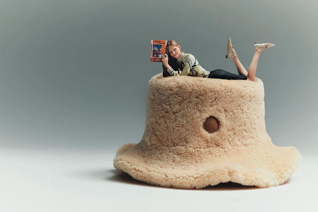 Woman lies on a beige, furry mushroom-shaped ottoman, reading a book in a studio setting with soft lighting and a simple, clean background.