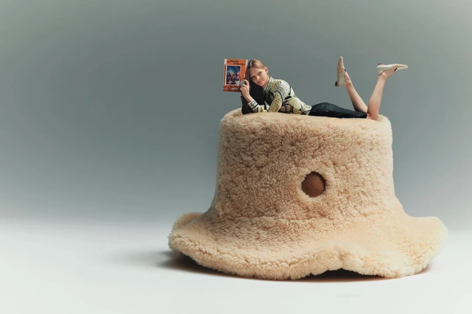 Woman lies on a beige, furry mushroom-shaped ottoman, reading a book in a studio setting with soft lighting and a simple, clean background.