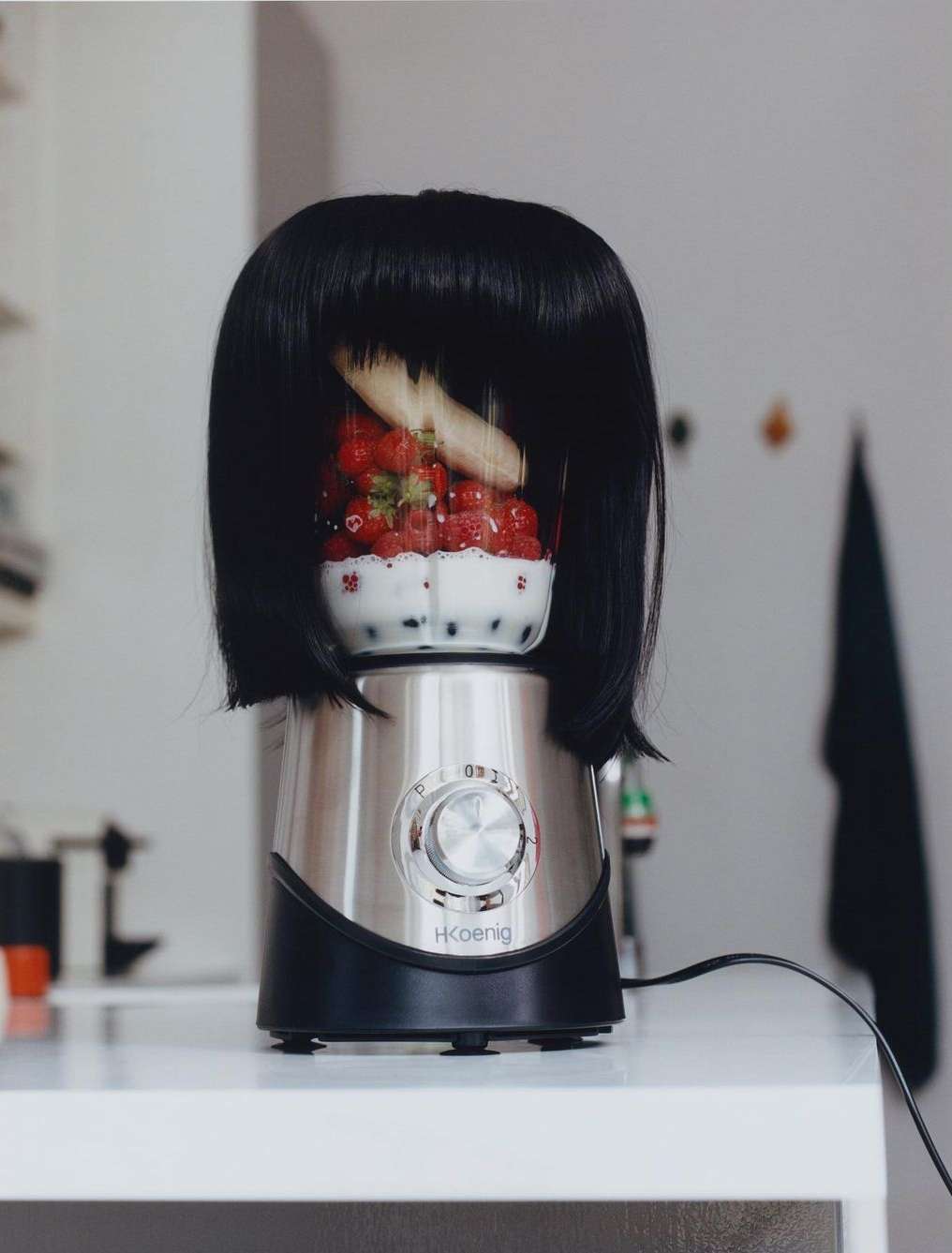 A quirky still life of a blender with a black wig, filled with strawberries and a banana, sitting on a white countertop in a kitchen setting.