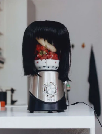 A quirky still life of a blender with a black wig, filled with strawberries and a banana, sitting on a white countertop in a kitchen setting.