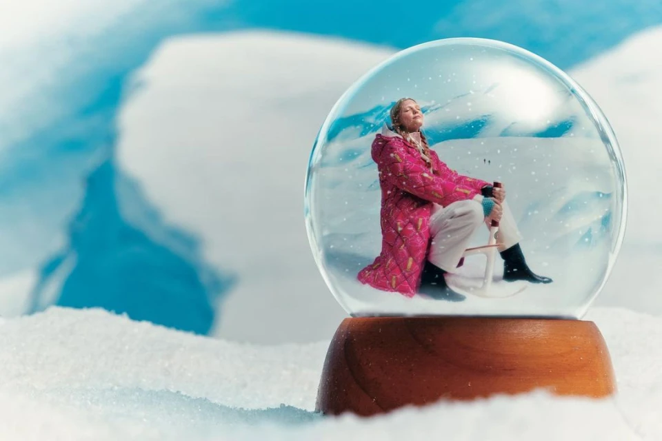A blonde woman in a pink coat sits in a snow globe with snowy mountains in the background, conveying a whimsical winter scene.