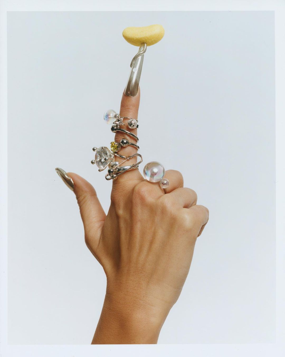 Woman's hand displaying rings on fingers with long silver nails, holding yellow blob on one finger, set against white background.