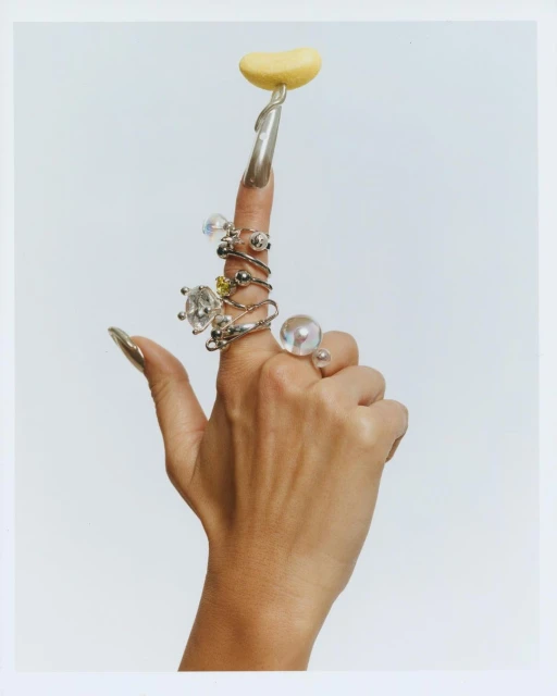 Woman's hand displaying rings on fingers with long silver nails, holding yellow blob on one finger, set against white background.