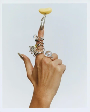 Woman's hand displaying rings on fingers with long silver nails, holding yellow blob on one finger, set against white background.