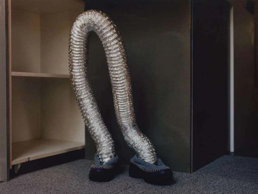 A silver flexible duct with shoes bends and appears to walk inside an empty shelving unit against a neutral backdrop.