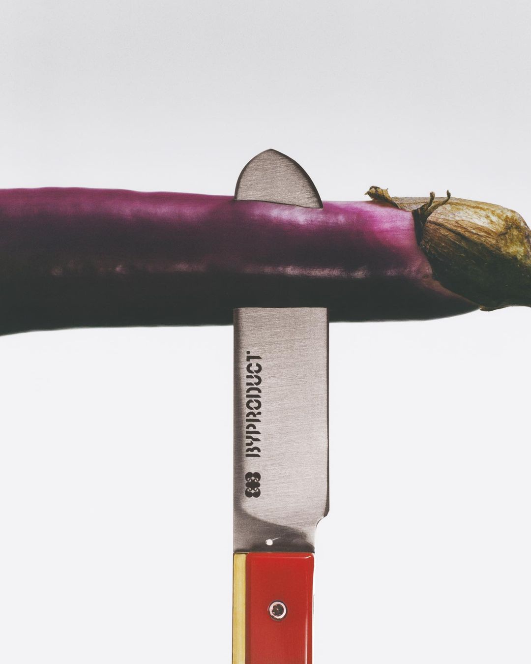 Studio shot of a purple eggplant sliced by a knife with a red handle against a white background with soft studio lighting.
