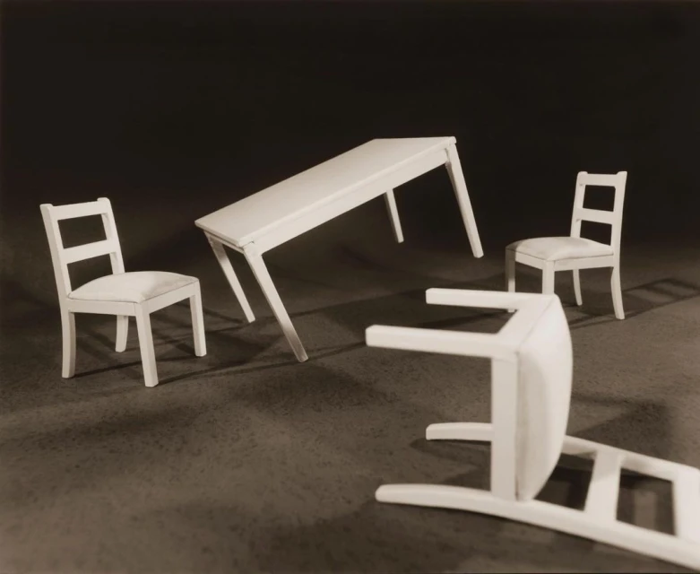 Still life of white table and chairs on a dark background, appearing to float in the air, casting shadows, creating a surreal composition.