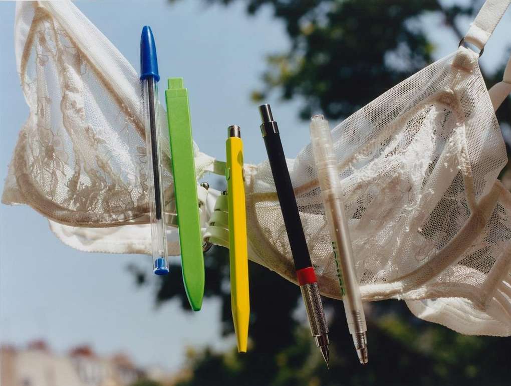 A white lace bra holds an array of colorful pens suspended in front of a blurred background of trees and rooftops in daylight.