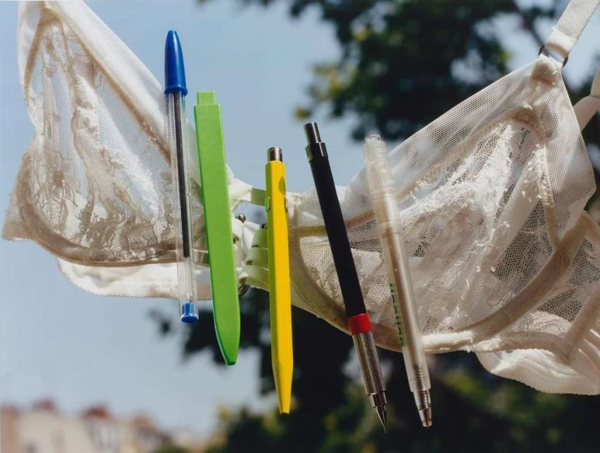 A white lace bra holds an array of colorful pens suspended in front of a blurred background of trees and rooftops in daylight.