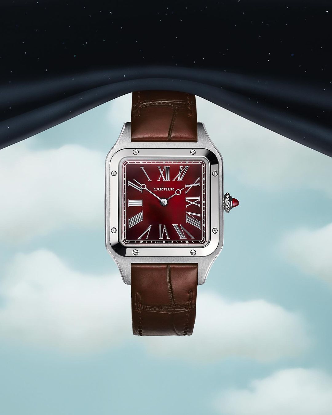 Luxury silver Cartier Santos watch with a red face and brown leather strap against a dreamy blue and black cloudy sky background.