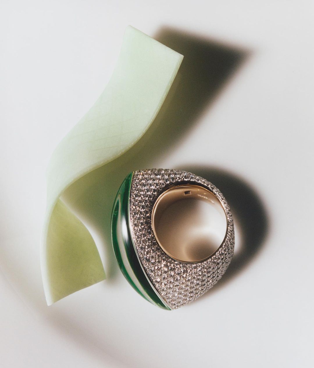 A unique green and gold ring is displayed with a pale green ribbon accent on a plain white surface in a product shot.
