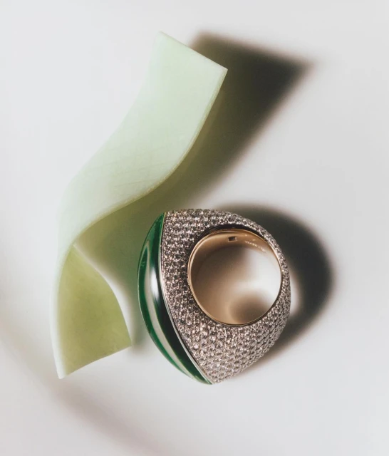 A unique green and gold ring is displayed with a pale green ribbon accent on a plain white surface in a product shot.