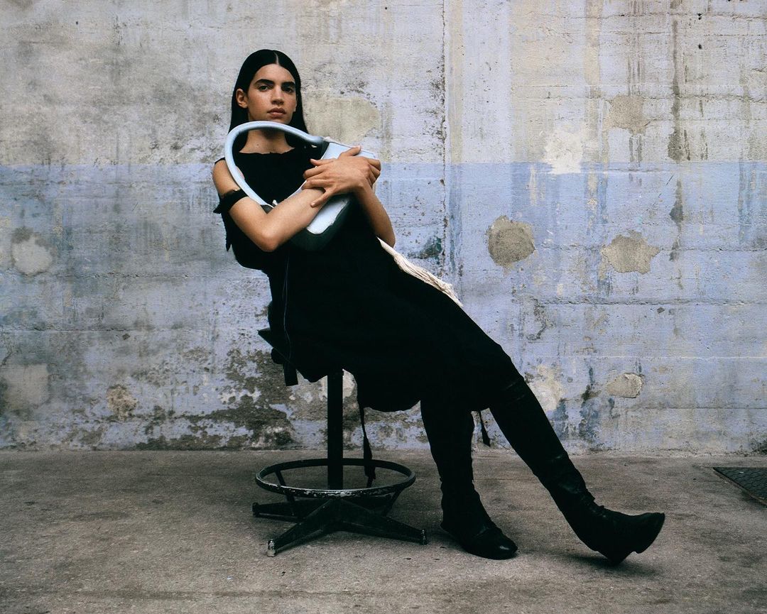 Young woman in a black dress and boots sits on a metal stool holding a light blue handbag against a concrete wall in a studio.