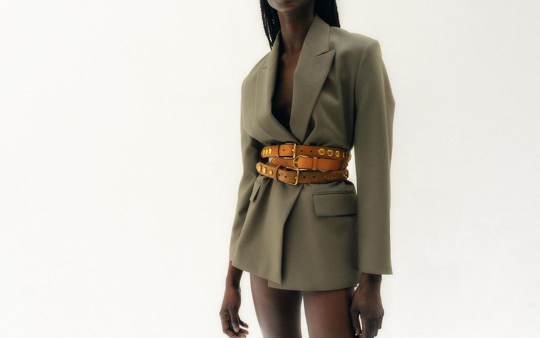 An African American woman wearing a tan blazer with multiple brown leather belts and brass hardware against a white backdrop.