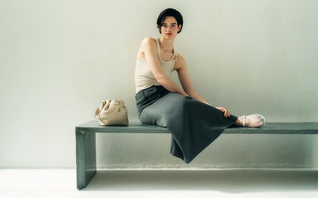 Woman in beige tank top and dark skirt sits on a bench next to a handbag in front of a pale wall in an indoor setting.