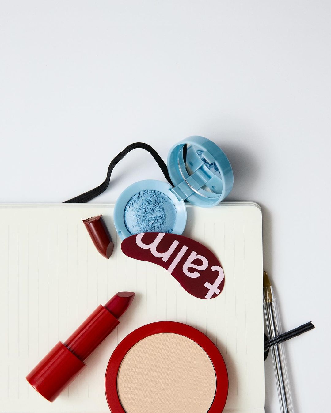 Still life composition featuring red lipstick, face powder, and blue eyeshadow on a white notebook, arranged against a white backdrop.
