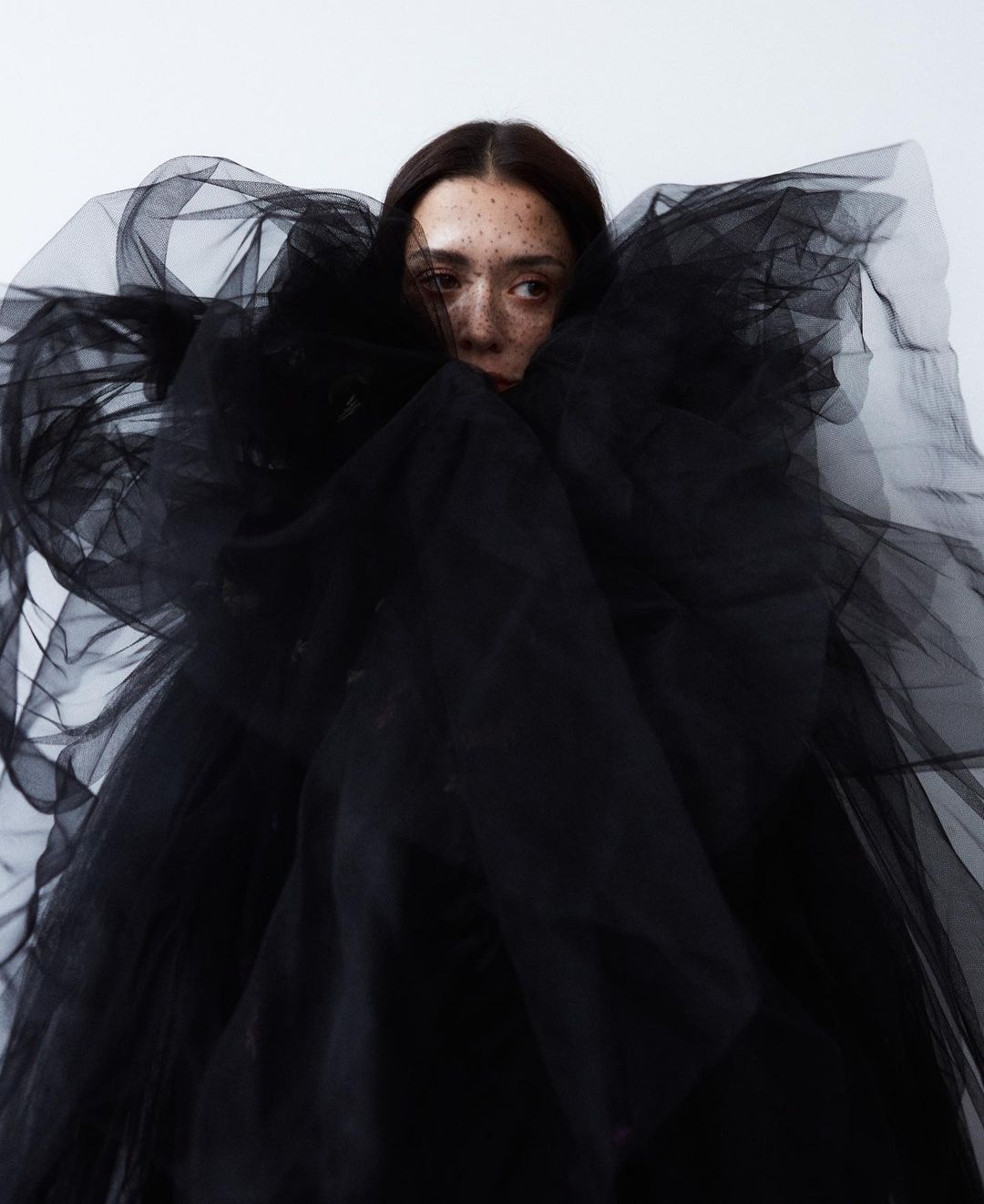 Portrait of a woman surrounded by a voluminous black tulle garment, set against a bright white backdrop.