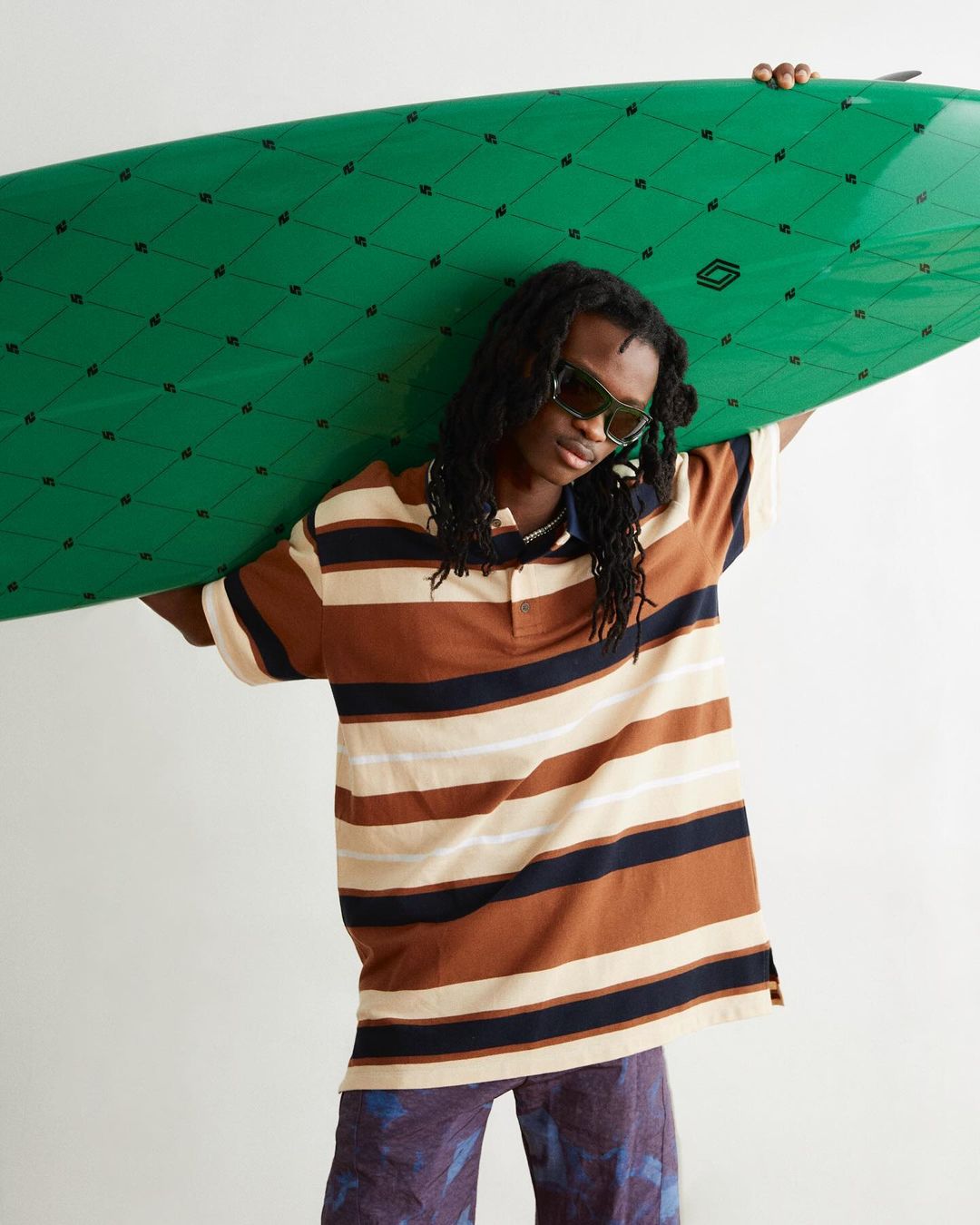 A stylish young adult with dreadlocks and sunglasses carries a green surfboard in a studio setting, wearing a striped brown polo shirt.