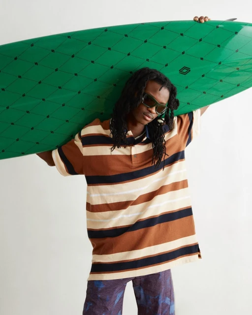 A stylish young adult with dreadlocks and sunglasses carries a green surfboard in a studio setting, wearing a striped brown polo shirt.