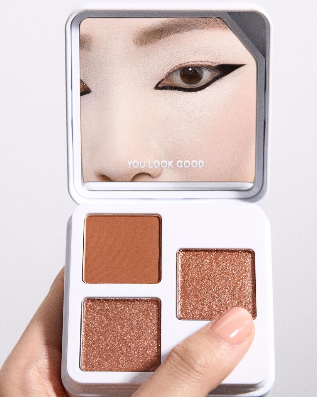 Eye shadow palette reflected in a mirror with shades of brown and gold, held by a fair-skinned hand with painted nails on a white background.