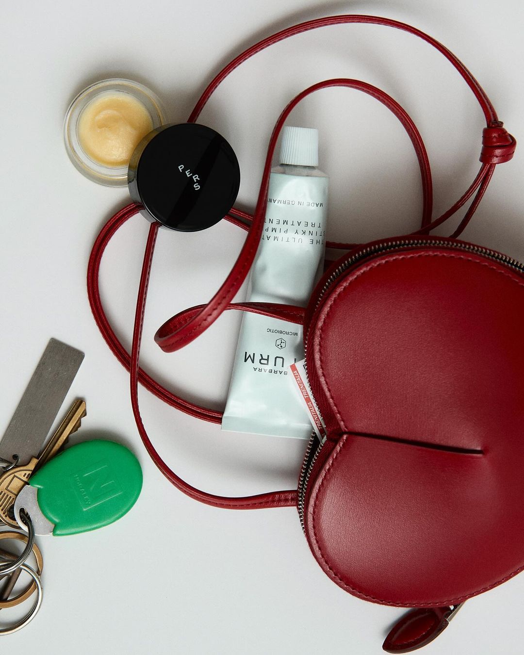 Overhead shot displays an open red purse, keys, and beauty products on a white surface, showcasing an organized, minimal composition.
