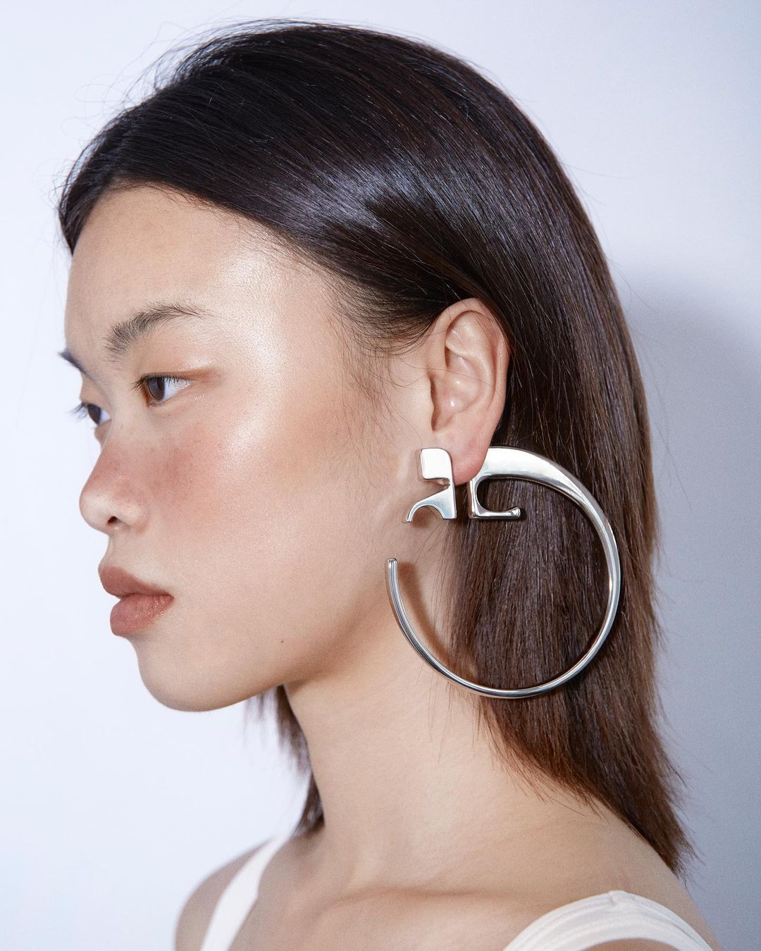Profile shot of a woman with dark hair wearing large silver earrings in a studio setting with a neutral background.