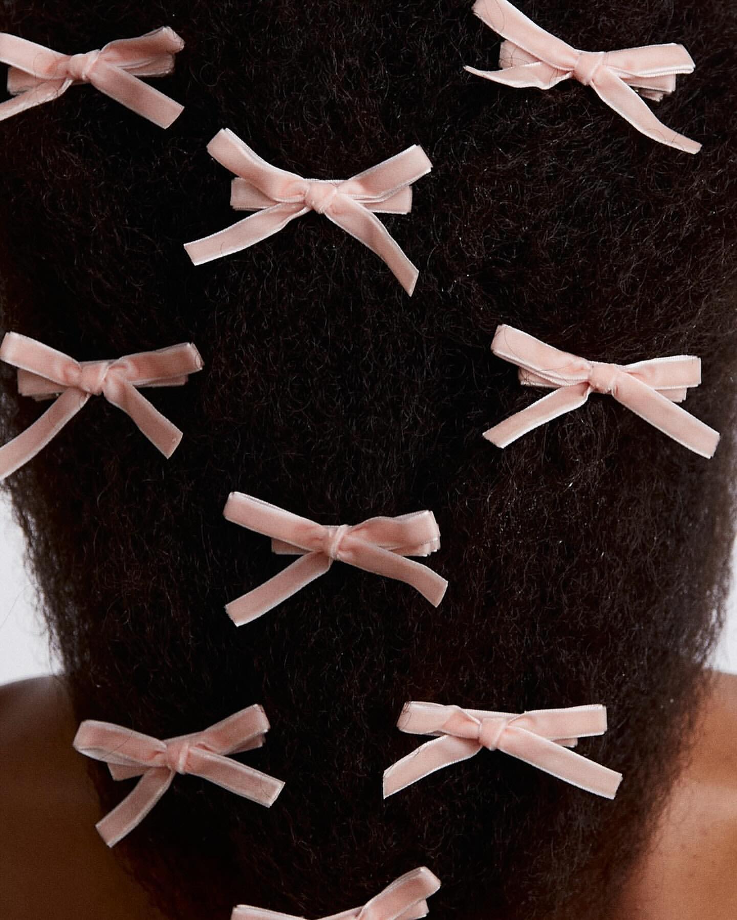 Close-up of a person's dark hair styled with multiple light pink, velvet, bow-shaped hair clips, set against a bright white background.