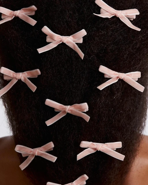 Close-up of a person's dark hair styled with multiple light pink, velvet, bow-shaped hair clips, set against a bright white background.
