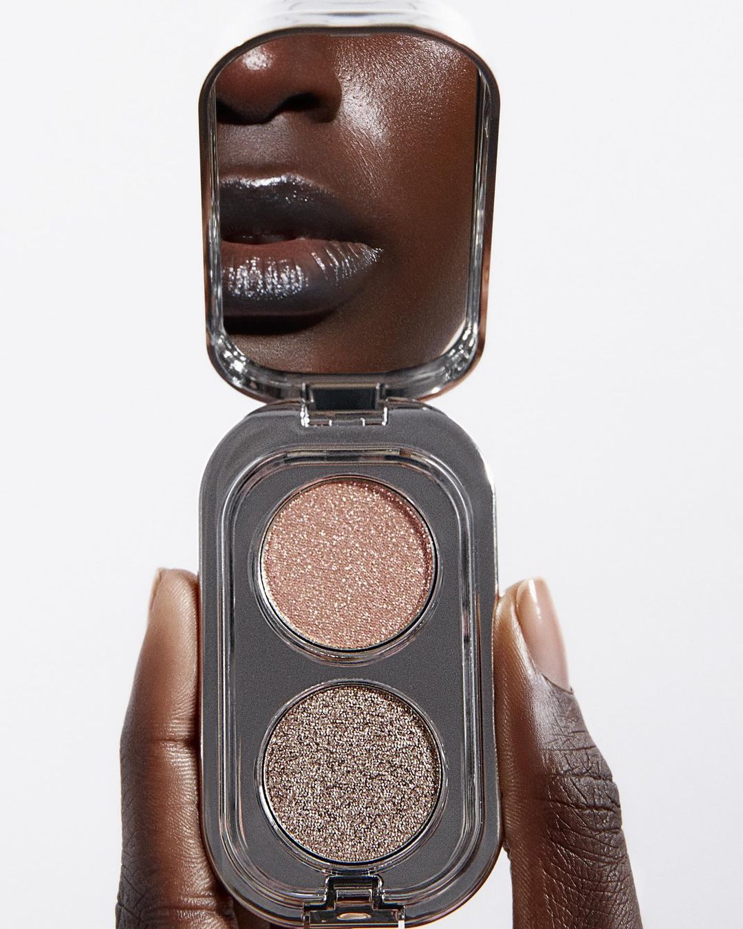 Close-up of an eyeshadow palette held in a dark-skinned hand, reflecting a person's face in the palette's mirror, against a bright white background.