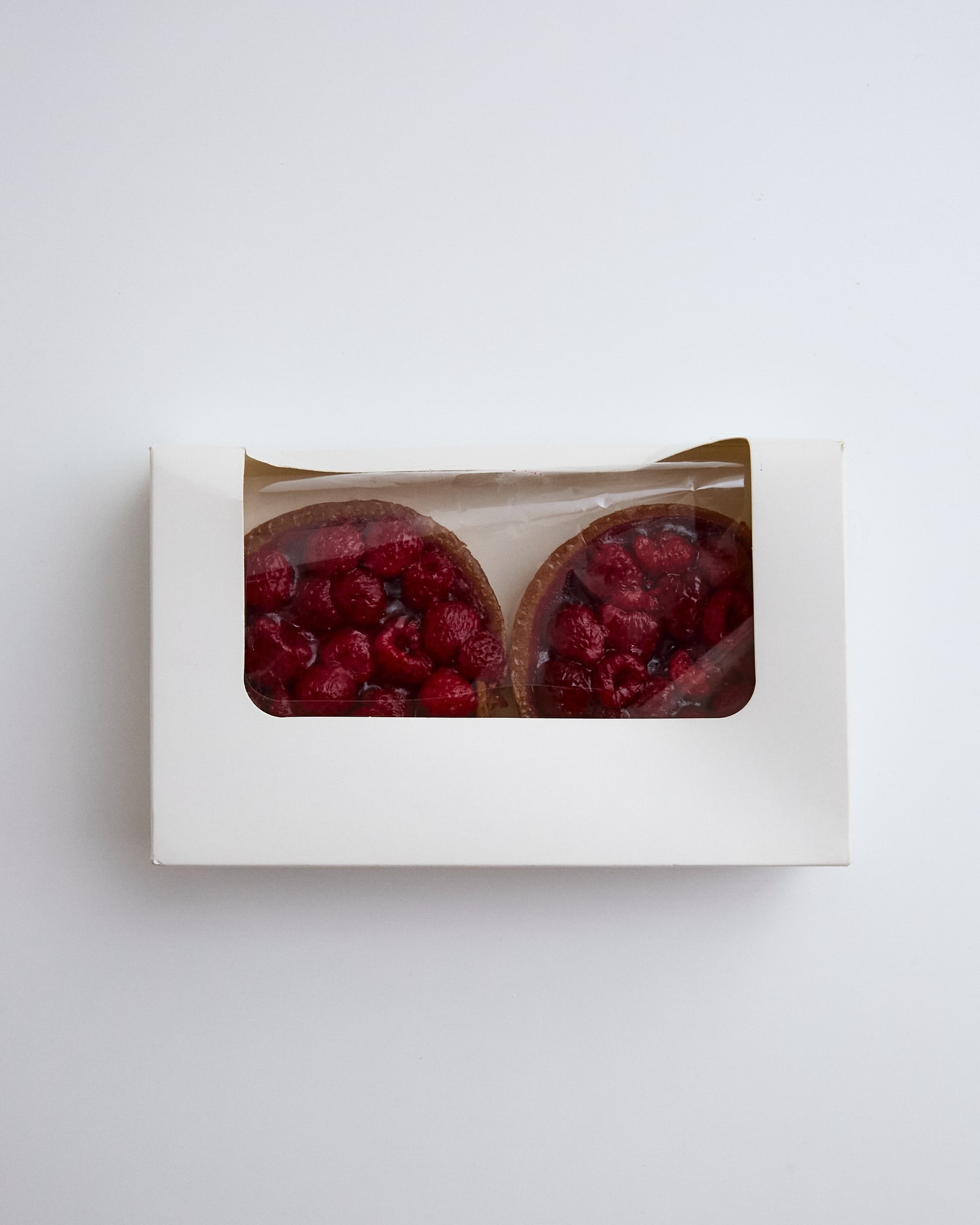Two raspberry tarts in a white cardboard box, viewed from above, on a white background, showcasing fresh pastry treats.