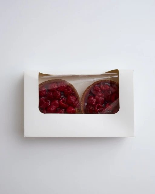 Two raspberry tarts in a white cardboard box, viewed from above, on a white background, showcasing fresh pastry treats.