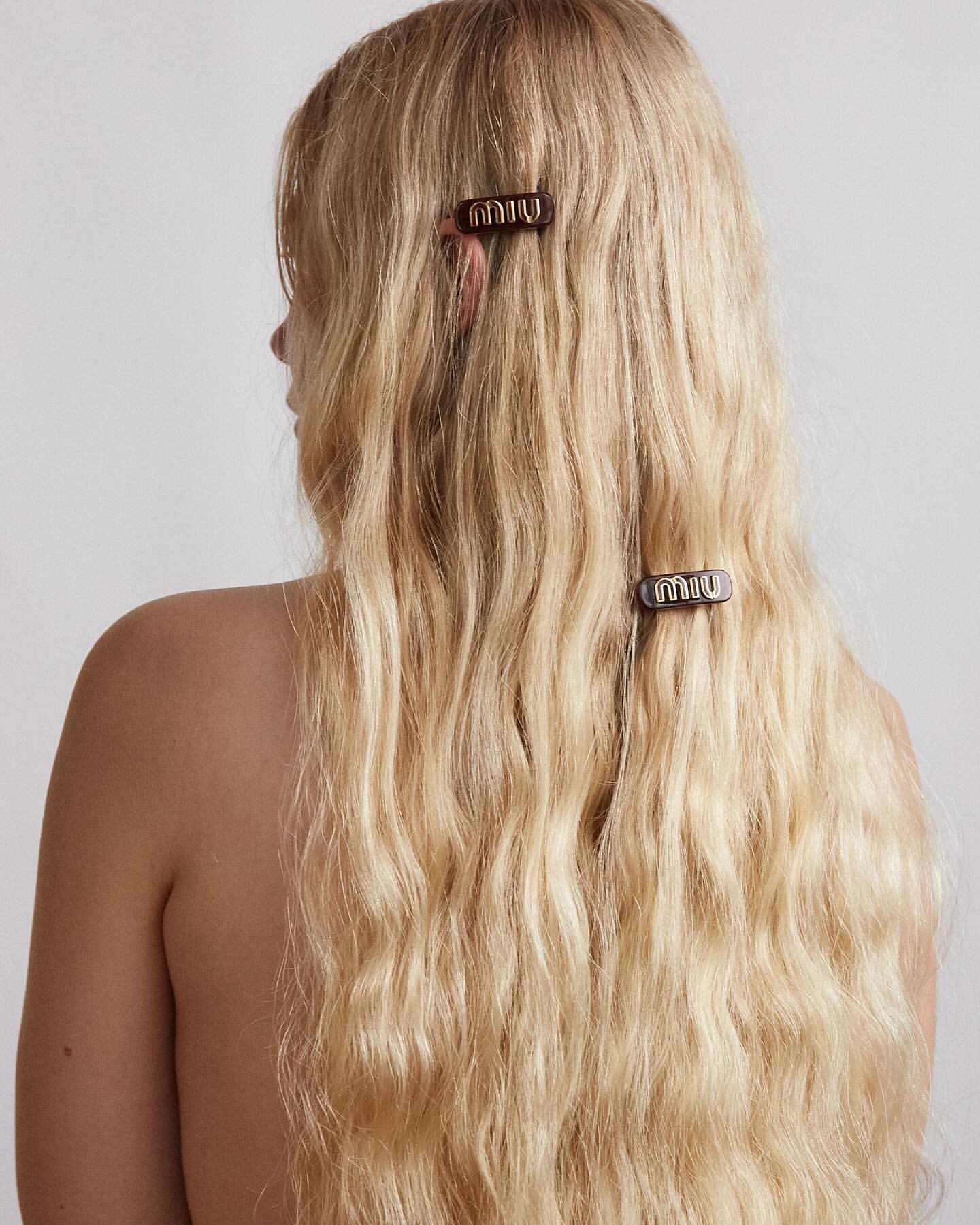 Woman's blonde hair styled with two Miu Miu logo hair clips against a white background, showcased from the back.