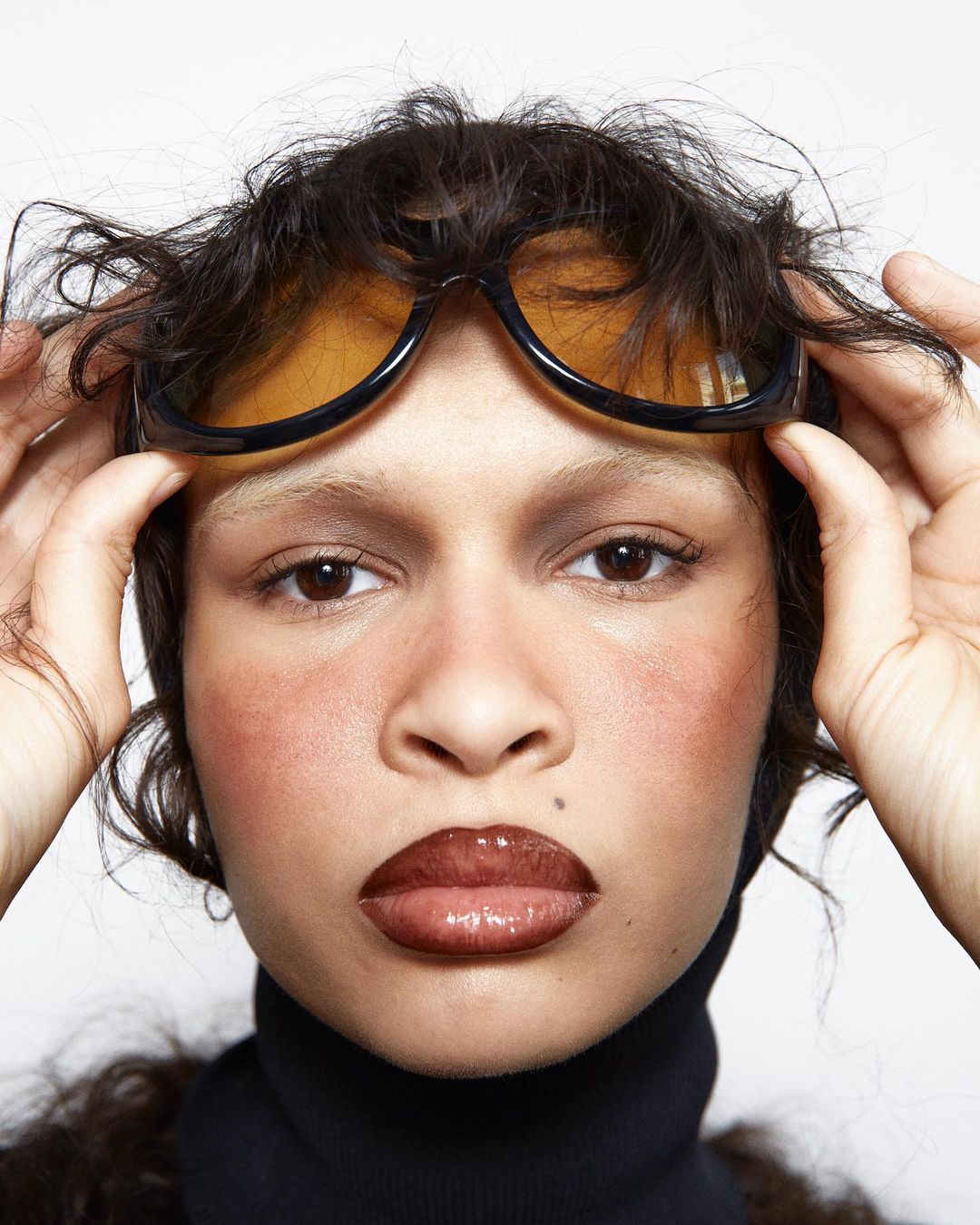 Close-up of an adult woman with dark hair and glossy lips, wearing yellow-tinted sunglasses and a black turtleneck against a white backdrop.