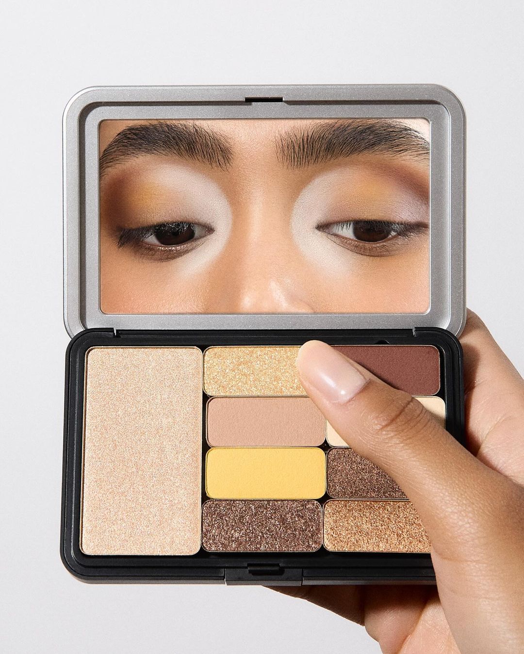 A hand holds a makeup palette with a mirror showing a close-up of a person's eyes. Palette features various colors, set against a plain background.