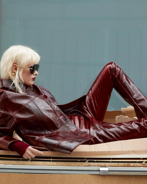 Fashion shot of a woman with sunglasses and a red vinyl outfit, lying in a cardboard box with a light blue background.