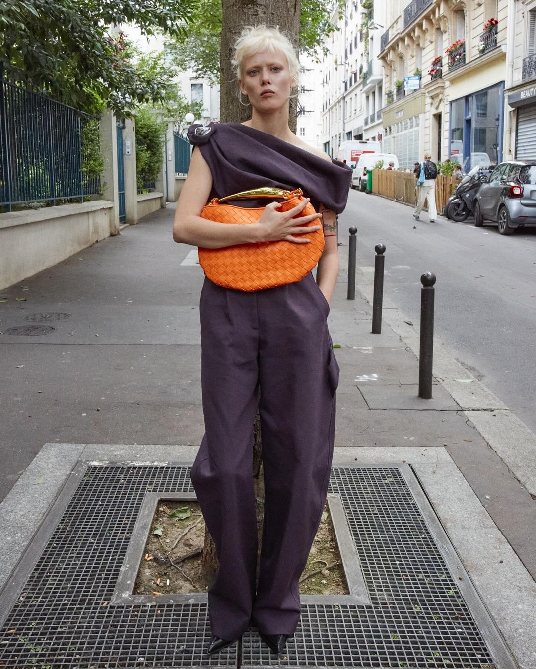 Woman in stylish purple jumpsuit stands outdoors with an orange woven handbag, posing against a tree on a city street in an editorial fashion shot.